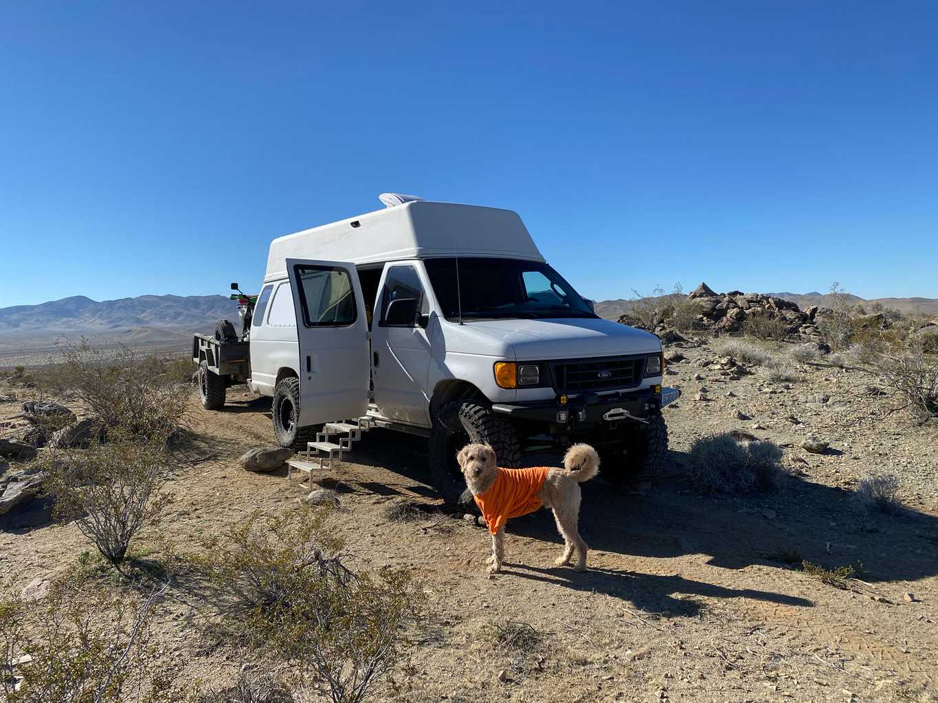 Our van and motorcycle trailer parked in the desert in California, with the clamshell door open and stairs out. Rainier the labradoodle is in the foreground in his orange t-shirt.